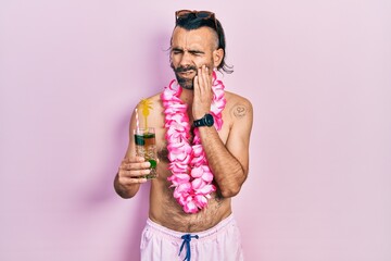 Young hispanic man wearing swimsuit and hawaiian lei drinking tropical cocktail touching mouth with...