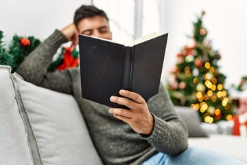 Fototapeta premium Young hispanic man reading book sitting on sofa by christmas tree at home