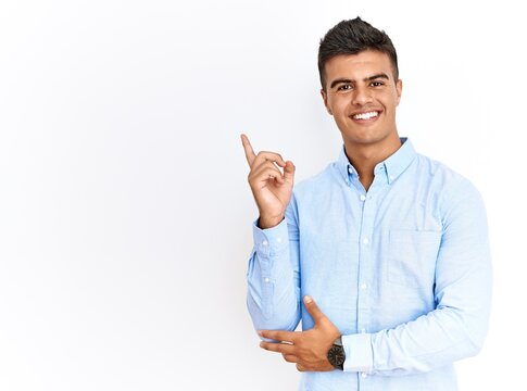 Young hispanic man wearing business shirt standing over isolated background with a big smile on face, pointing with hand finger to the side looking at the camera.