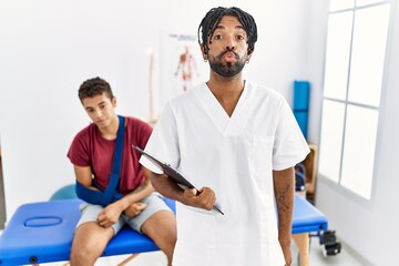 Young hispanic man working at pain recovery clinic with a man with broken arm looking at the camera blowing a kiss on air being lovely and sexy. love expression.