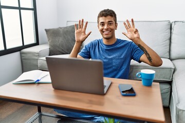 Young handsome hispanic man using laptop sitting on the floor showing and pointing up with fingers number nine while smiling confident and happy.