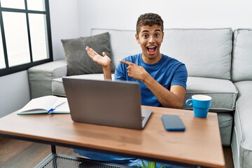 Young handsome hispanic man using laptop sitting on the floor amazed and smiling to the camera while presenting with hand and pointing with finger.
