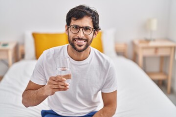 Young hispanic man drinking glass of water sitting on bed at bedroom