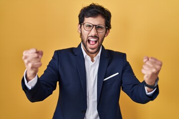 Handsome latin man standing over yellow background angry and mad raising fists frustrated and furious while shouting with anger. rage and aggressive concept.