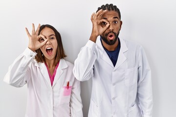 Young hispanic doctors standing over white background doing ok gesture shocked with surprised face, eye looking through fingers. unbelieving expression.