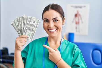 Young hispanic woman holding dollars banknotes working at pain recovery clinic smiling happy...