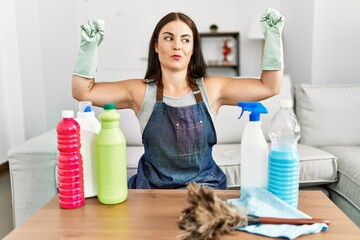 Young brunette woman wearing cleaner apron and gloves cleaning at home showing arms muscles smiling proud. fitness concept.