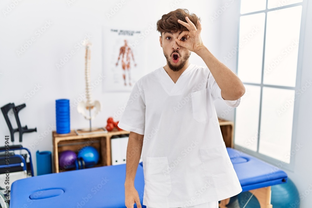 Poster young arab man working at pain recovery clinic doing ok gesture shocked with surprised face, eye loo