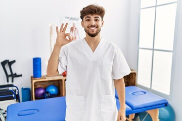 Obraz premium Young arab man working at pain recovery clinic smiling positive doing ok sign with hand and fingers. successful expression.
