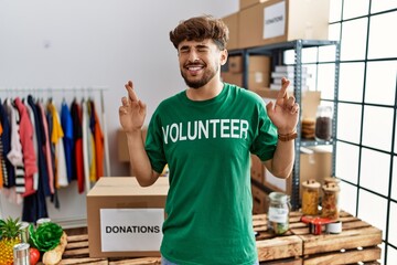 Young arab man wearing volunteer t shirt at donations stand gesturing finger crossed smiling with...