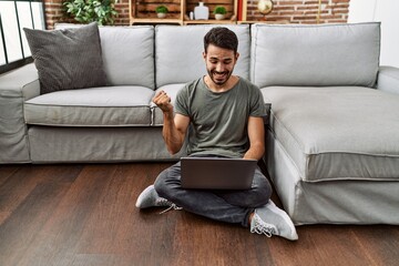 Young hispanic man using laptop sitting on the at home screaming proud, celebrating victory and success very excited with raised arm