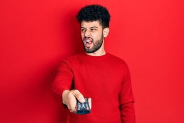 Young arab man with beard holding television remote control angry and mad screaming frustrated and...