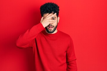 Young arab man with beard wearing casual red sweater peeking in shock covering face and eyes with hand, looking through fingers with embarrassed expression.