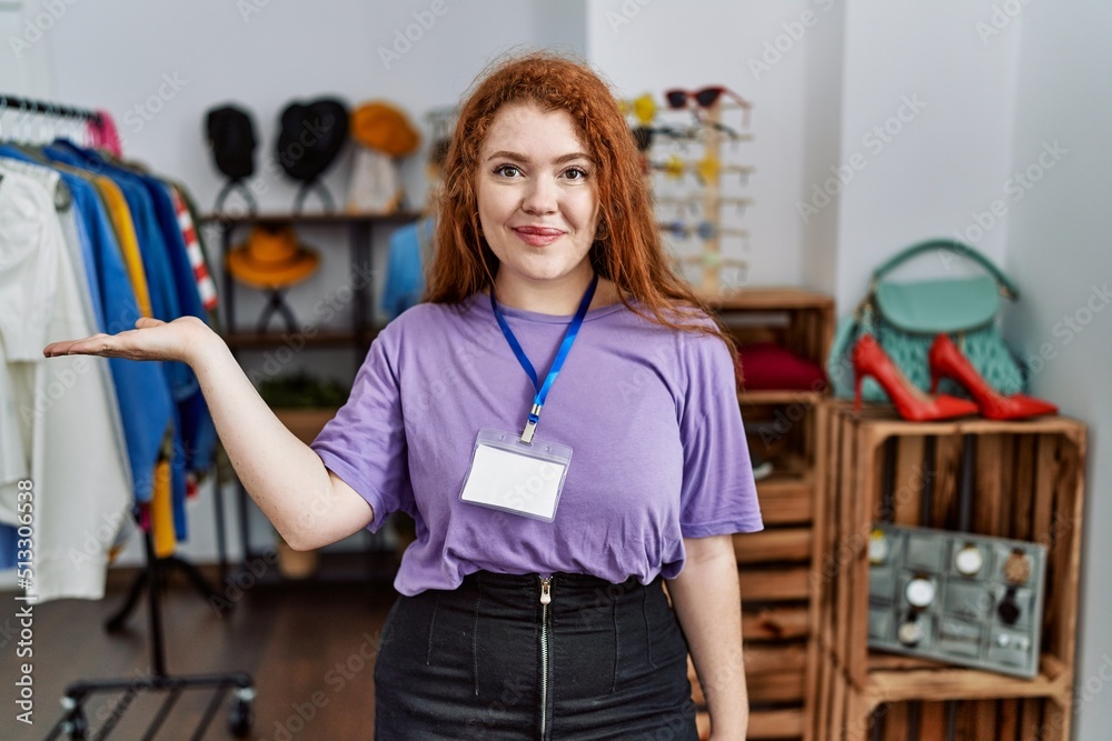 Wall mural Young redhead woman working as manager at retail boutique smiling cheerful presenting and pointing with palm of hand looking at the camera.