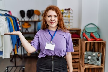 Young redhead woman working as manager at retail boutique smiling cheerful presenting and pointing with palm of hand looking at the camera.
