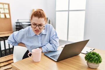 Young redhead woman working at the office using computer laptop suffering of backache, touching back with hand, muscular pain