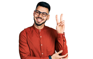 Young hispanic man with beard wearing business shirt and glasses smiling with happy face winking at...