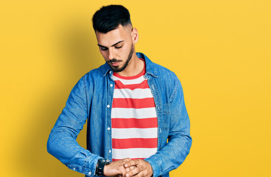 Young hispanic man with beard wearing casual denim jacket checking the time on wrist watch, relaxed and confident
