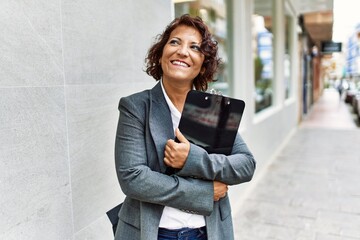 Middle age latin businesswoman smiling happy holding clipboard at the city. - obrazy, fototapety, plakaty