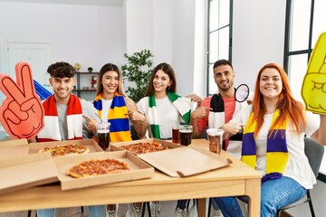 Group of young hispanic people eating pizza supporting soccer team at home smiling happy pointing...