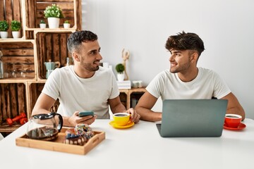 Two hispanic men couple having breakfast using smartphone and laptop at home