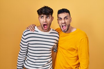 Young hispanic gay couple standing over yellow background angry and mad screaming frustrated and furious, shouting with anger. rage and aggressive concept.