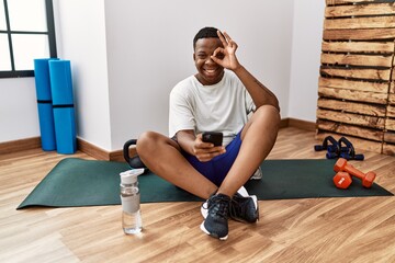 Young african man sitting on training mat at the gym using smartphone doing ok gesture with hand...