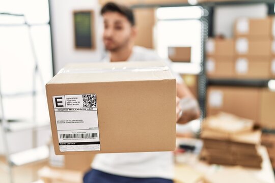 Young hispanic man working at small business ecommerce holding box smiling looking to the side and staring away thinking.