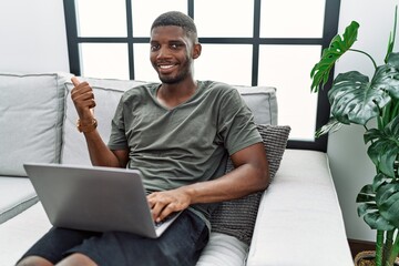 Young african american man using laptop at home sitting on the sofa pointing to the back behind with hand and thumbs up, smiling confident