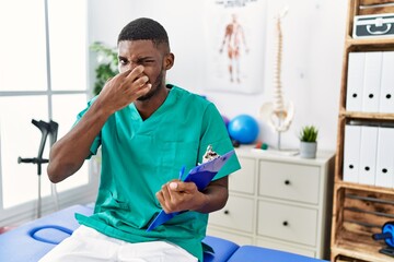 Young african american man working at pain recovery clinic smelling something stinky and...