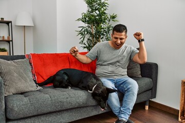 Young latin man and dog sitting on the sofa at home dancing happy and cheerful, smiling moving...