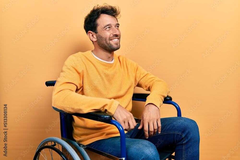 Poster Handsome man with beard sitting on wheelchair looking away to side with smile on face, natural expression. laughing confident.