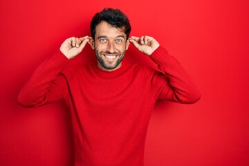 Handsome man with beard wearing casual red sweater smiling pulling ears with fingers, funny...