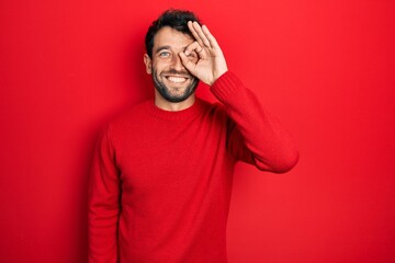 Handsome man with beard wearing casual red sweater doing ok gesture with hand smiling, eye looking through fingers with happy face. © Krakenimages.com