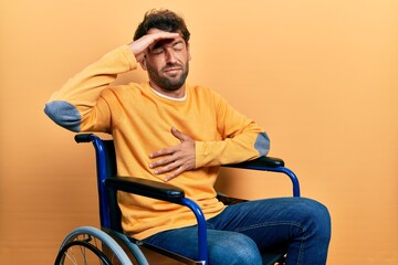 Handsome man with beard sitting on wheelchair touching forehead for illness and fever, flu and cold, virus sick