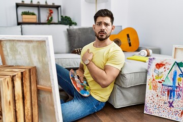 Young man with beard painting canvas at home pointing aside worried and nervous with forefinger,...
