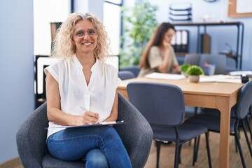 Middle age woman working at the office holding clipboard looking positive and happy standing and smiling with a confident smile showing teeth