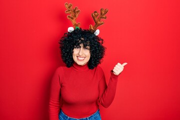 Young middle east woman wearing cute christmas reindeer horns smiling with happy face looking and pointing to the side with thumb up.