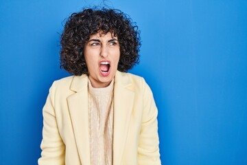 Fototapeta na wymiar Young brunette woman with curly hair standing over blue background angry and mad screaming frustrated and furious, shouting with anger. rage and aggressive concept.