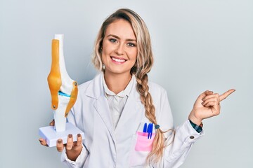 Beautiful young blonde doctor woman holding anatomical model of knee joint smiling happy pointing...