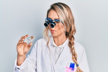 Beautiful young blonde woman holding brilliant diamond stone wearing magnifier glasses making fish...