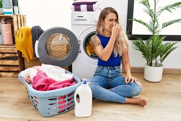 Young beautiful woman doing laundry sitting by wicker basket smelling something stinky and disgusting, intolerable smell, holding breath with fingers on nose. bad smell