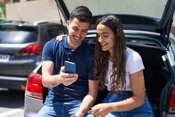 Fototapeta na wymiar Young hispanic couple sitting on car trunk using smartphone at street