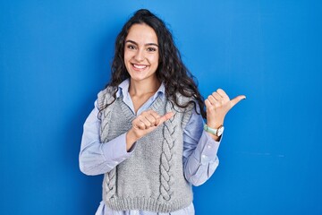 Young brunette woman standing over blue background pointing to the back behind with hand and thumbs up, smiling confident