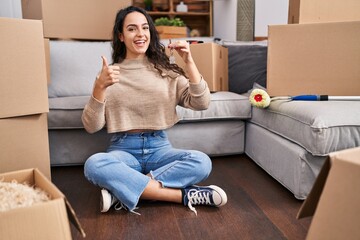 Young brunette woman sitting on the floor at new home holding keys smiling happy and positive, thumb up doing excellent and approval sign