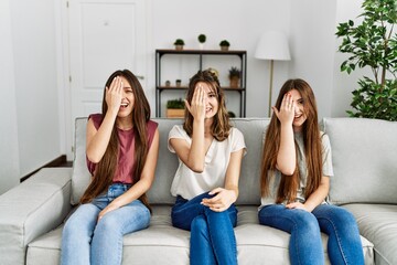 Group of three hispanic girls sitting on the sofa at home covering one eye with hand, confident smile on face and surprise emotion.