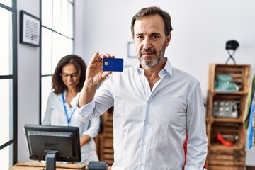 Hispanic man holding credit card at retail shop thinking attitude and sober expression looking self confident