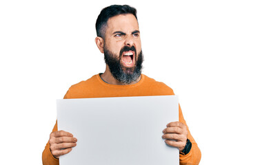 Hispanic man with beard holding blank empty banner angry and mad screaming frustrated and furious, shouting with anger. rage and aggressive concept.