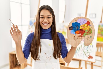 Young latin woman smiling confident holding paintbrush and palette at art studio