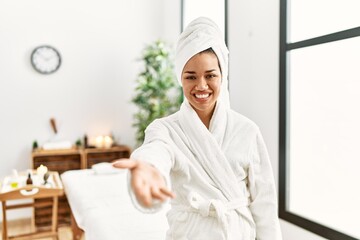 Young brunette woman wearing towel and bathrobe standing at beauty center smiling friendly offering...